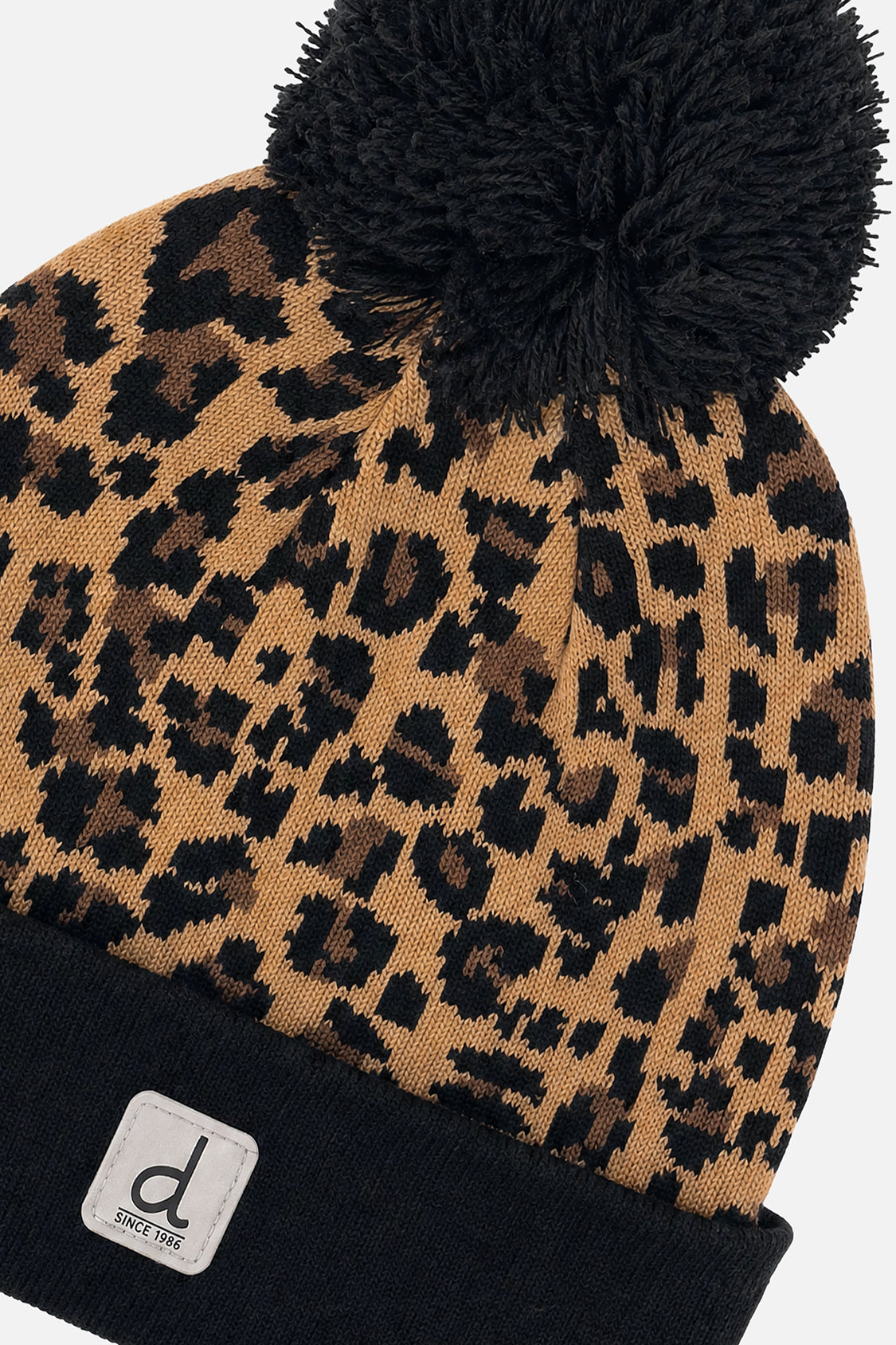Close-up of the brown leopard jacquard knit, black cuff, logo patch and black pompom on the Deux par Deux hat