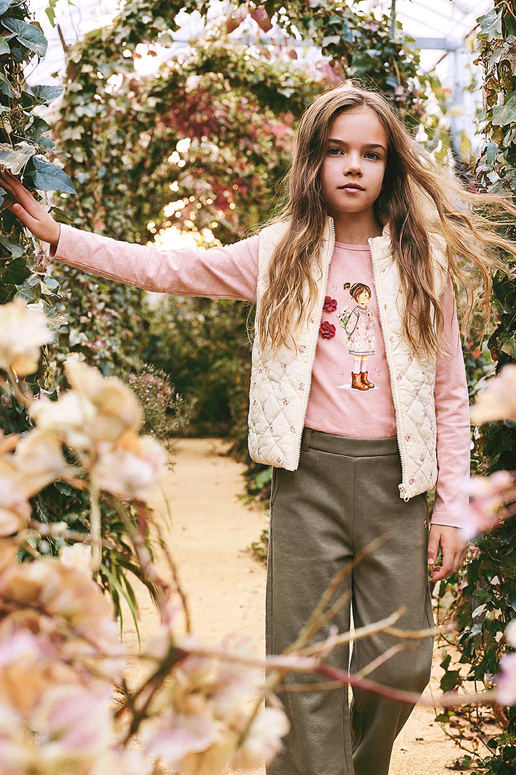 Girl in a pink top, cream quilted vest and sage green flare pants in a flower garden