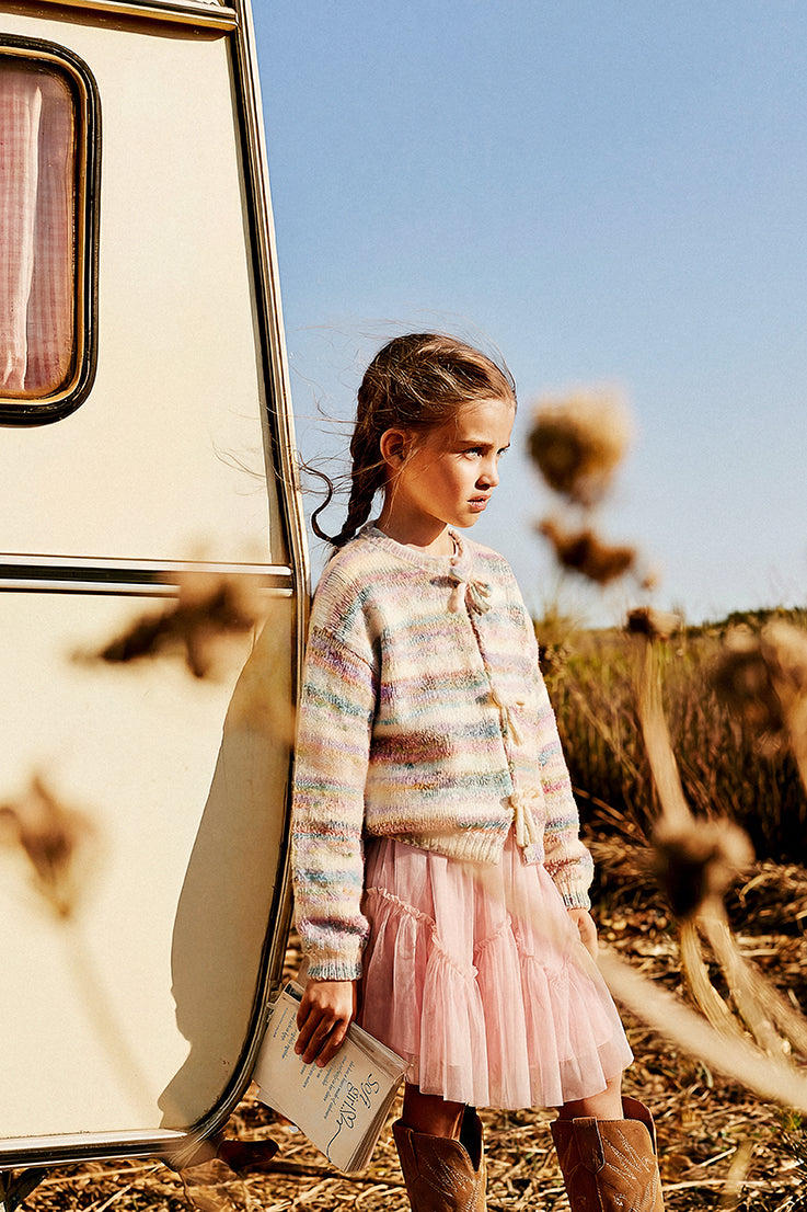Child in the pink mesh skirt and a striped cardigan beside a camper