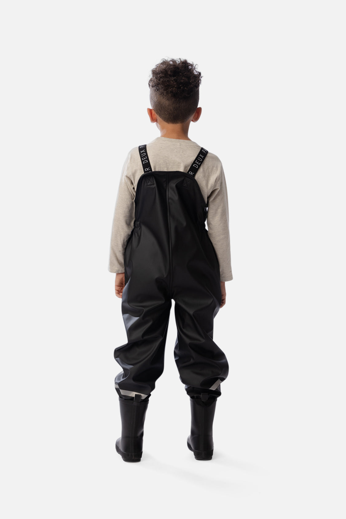 Back view of a child wearing the black rain overalls over a beige long-sleeve top
