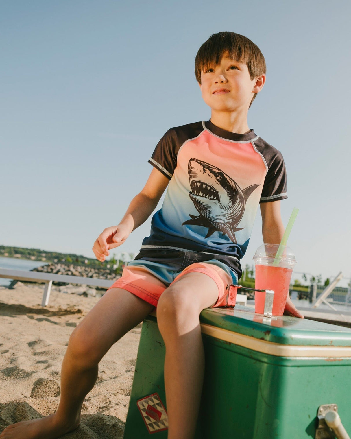 Boy sitting near the water wearing blue/orange gradient shark rashguard and matching trunks