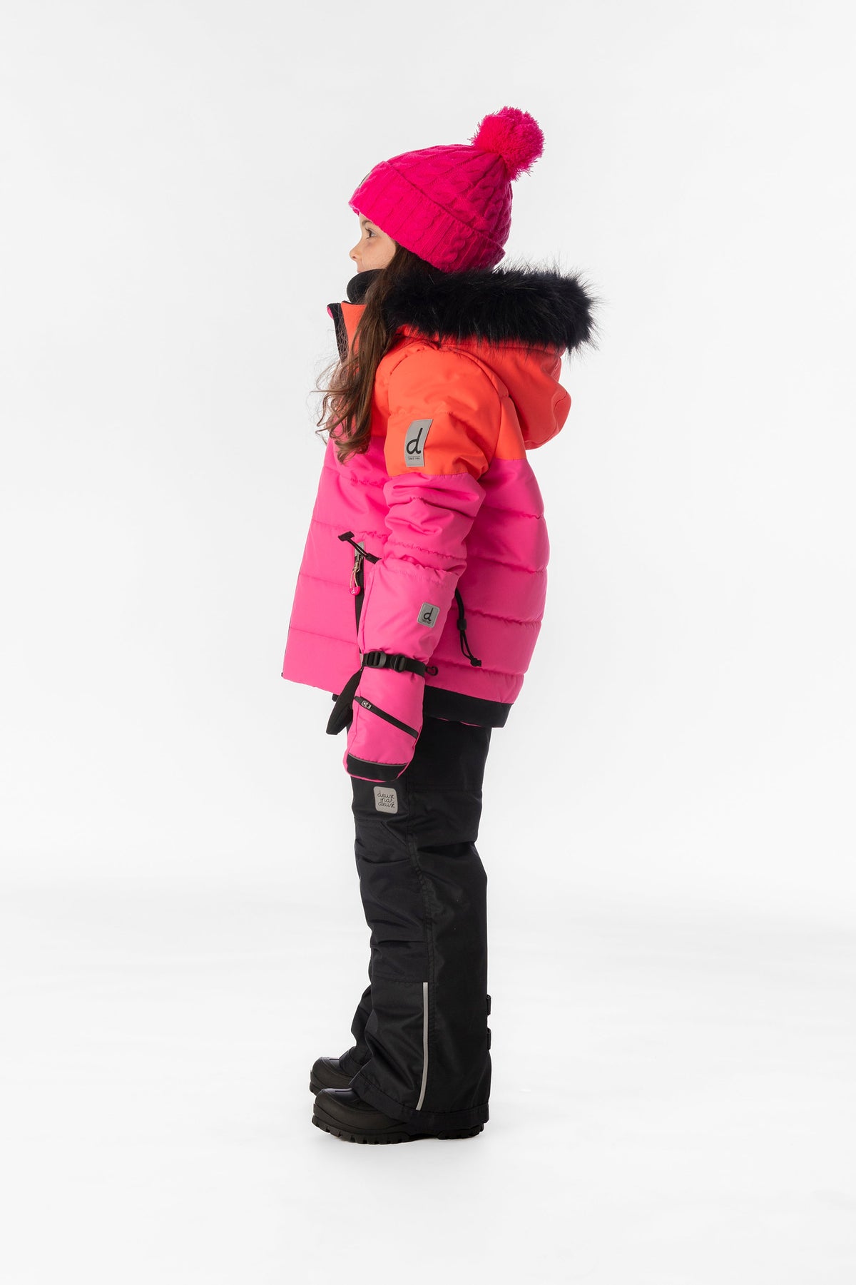 Girl in the coral, bright pink and black Deux par Deux PLAY snowsuit, side view