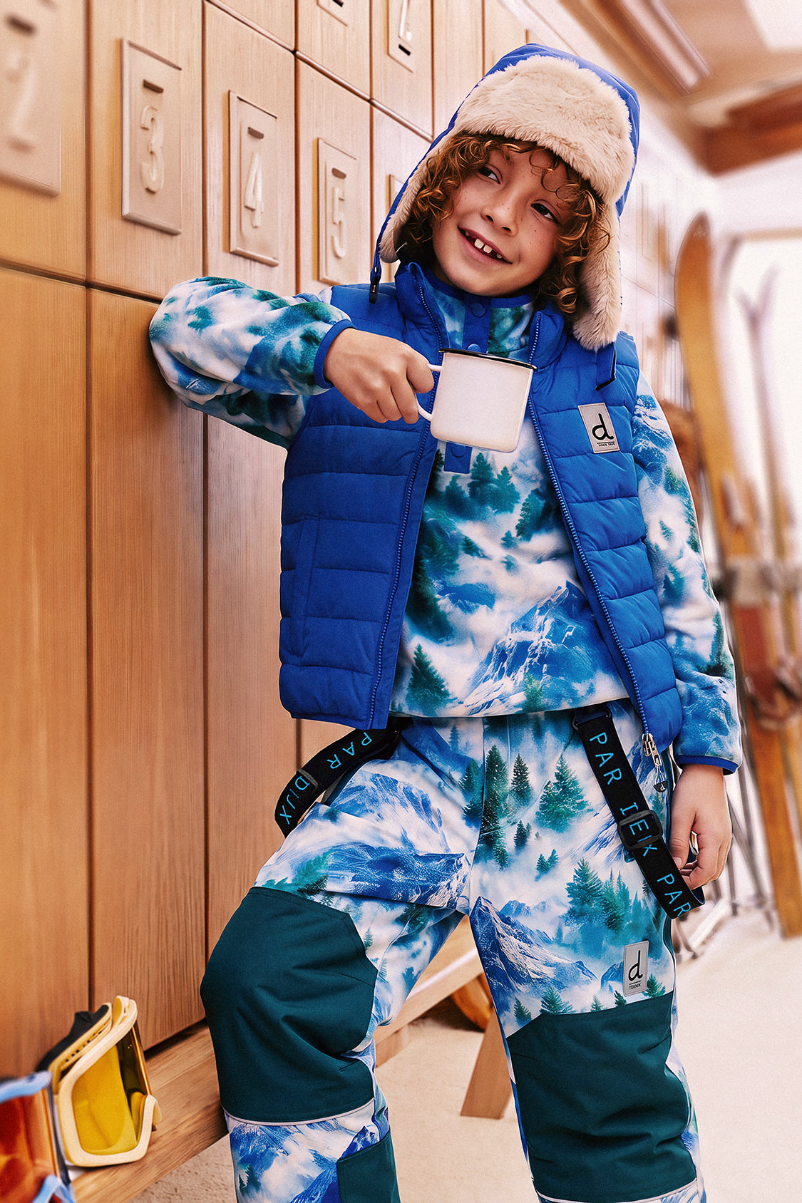 Child wearing the blue Deux par Deux lined chapka in a ski lodge with a blue puffer vest and mountain-print snow outfit