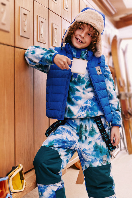 Child wearing the blue Deux par Deux lined chapka in a ski lodge with a blue puffer vest and mountain-print snow outfit