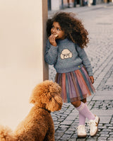 Child in a gray poodle sweater and the multicolor mesh skirt beside a poodle on a cobblestone street