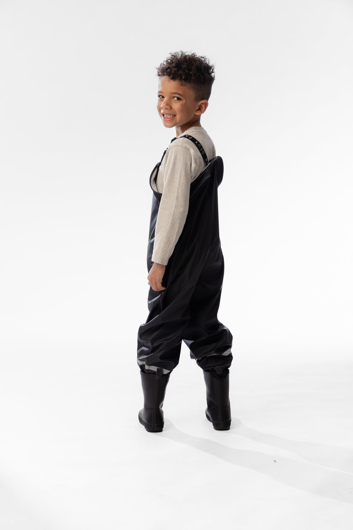 Side view of a child in the Deux par Deux black rain overalls and black rain boots